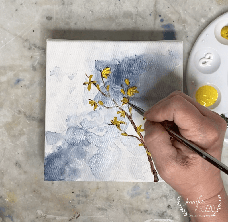 A hand demonstrates how to paint a forsythia branch with yellow flowers on a blue and white abstract background on a small canvas, with a paint palette nearby.
