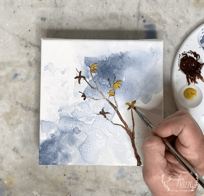 A hand demonstrates how to paint a forsythia branch, brushing yellow flowers onto a square canvas with watercolors, while a palette holding brown and yellow paint sits nearby.
