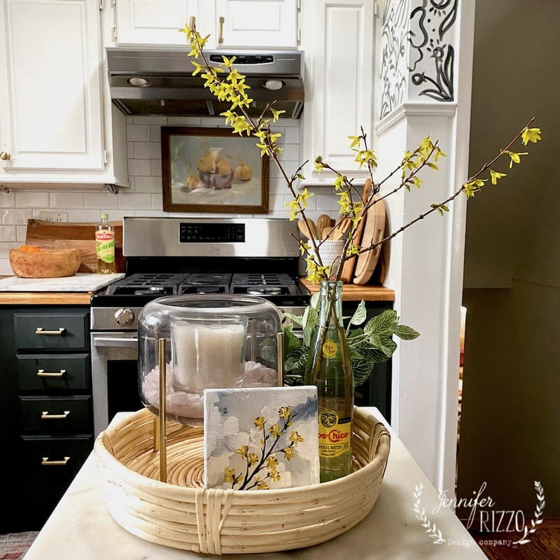 A kitchen counter displays a round wooden tray with a candle, floral artwork, and a glass bottle showcasing how to paint a forsythia branch in vivid yellow. In the background, there's a painting alongside the stove and cabinets.