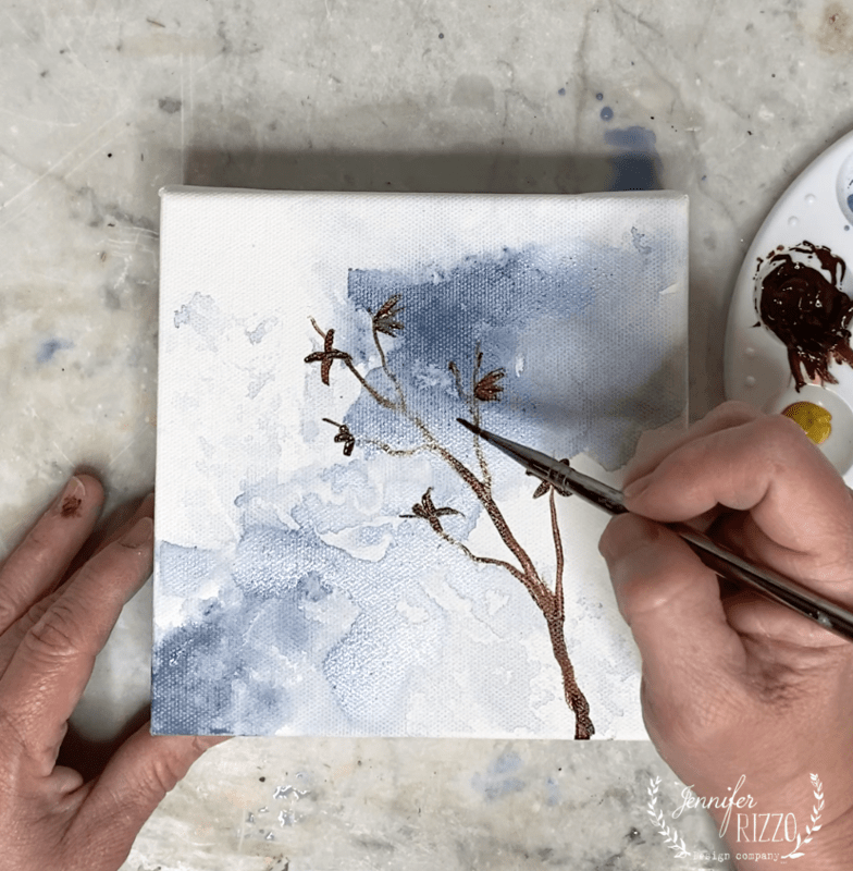 A hand demonstrates how to paint a forsythia branch with flowers on a small canvas with a blue and white background; a paint palette sits nearby.