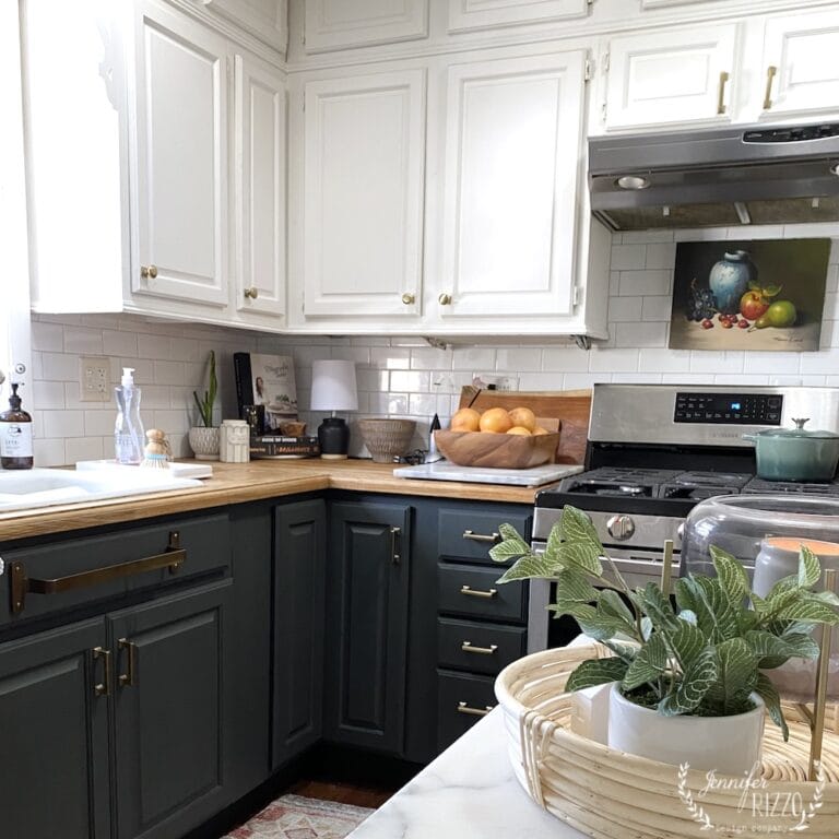 A kitchen with green and white kitchen cabinets, wooden countertops, a gas stove, and various kitchen items and plants on the counters.