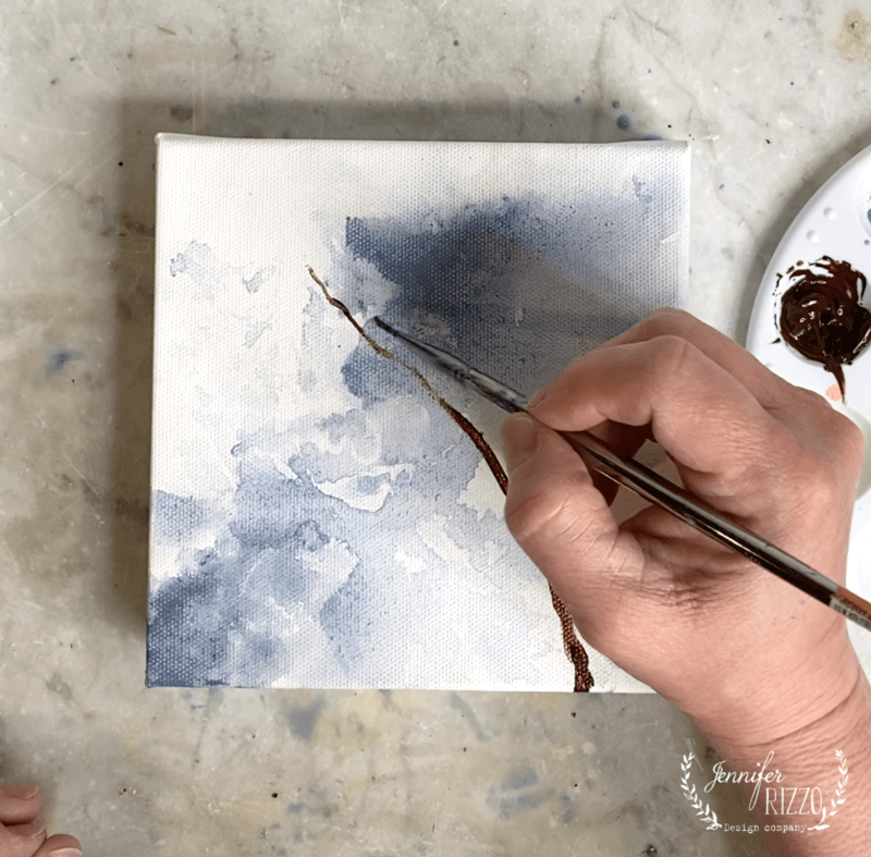 A hand uses a paintbrush to add a brown line, demonstrating how to paint a forsythia branch on an abstract blue and white canvas. A paint palette is visible in the corner.