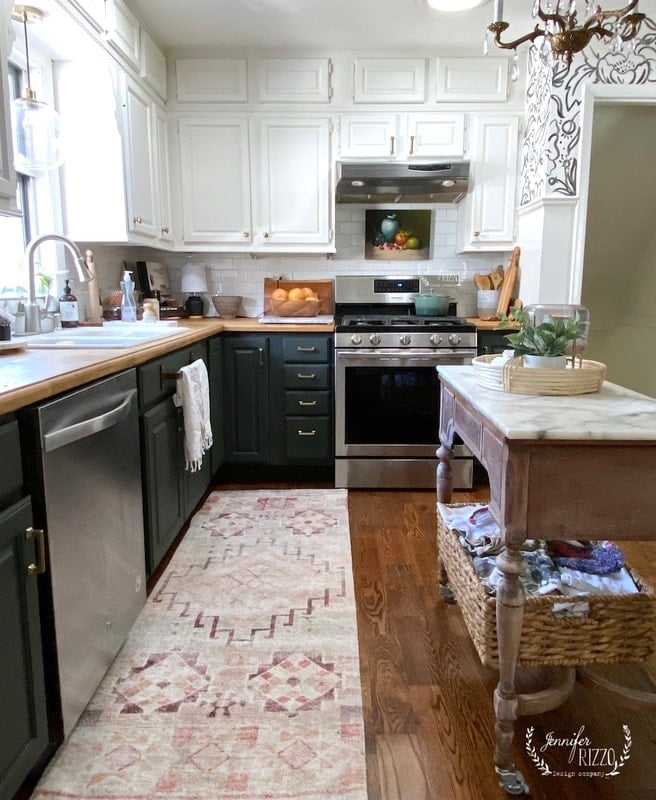 A kitchen with green and white cabinets, a stainless steel stove, farmhouse sink, wooden countertops, and a small island with a basket underneath on a patterned rug.