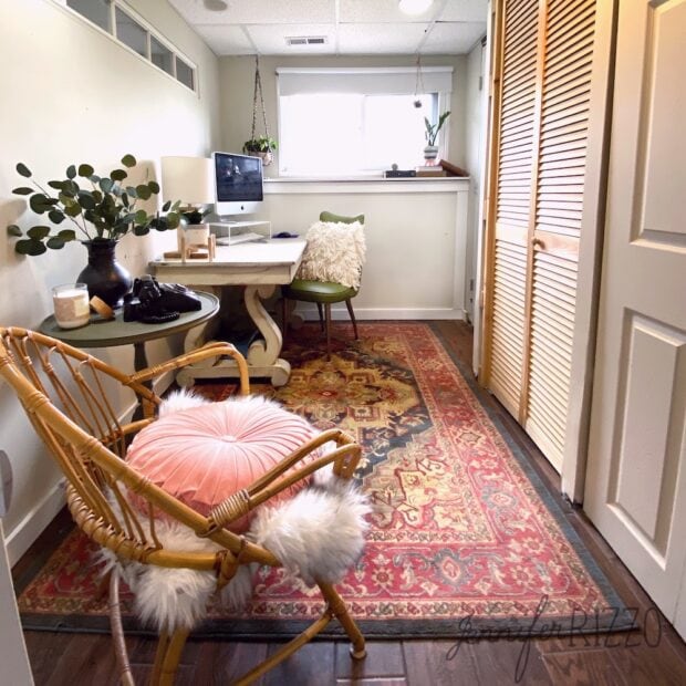 Home office with a computer on a white desk, green chair, rattan chair with pink cushion, decorative rug, and indoor plants. Light streams in from a window at the back in this no money spent office makeover.