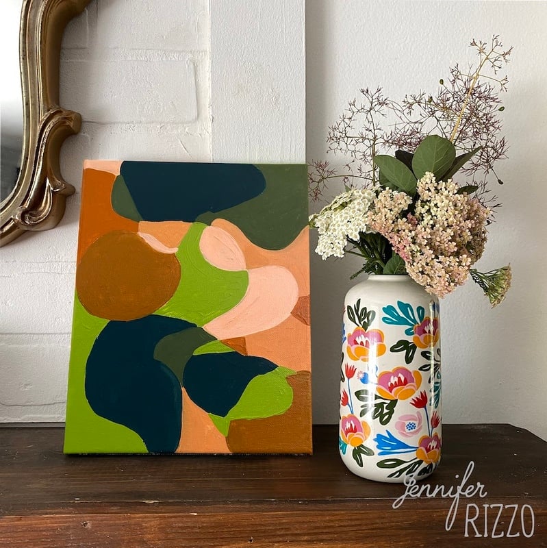 Abstract painting featuring easy line fill abstract art with green, peach, and blue shapes beside a white vase adorned with colorful floral designs, holding a bouquet of flowers on a dark wooden surface.