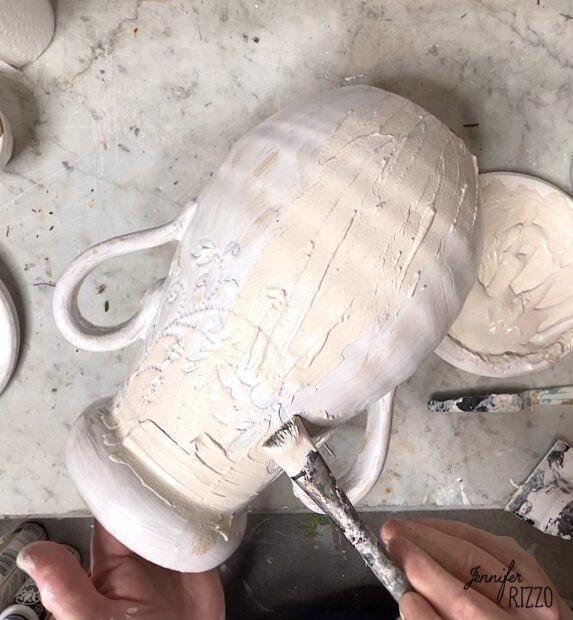 A person gives a thrift store vase a plaster-look makeover by applying off-white paint to the textured ceramic with a paintbrush, while a paint bowl and brush sit nearby on the work surface.