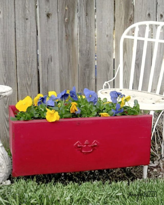 A repurposed dresser drawer into a planter holds yellow and purple flowers, set on grass beside a white metal chair and a wooden fence.