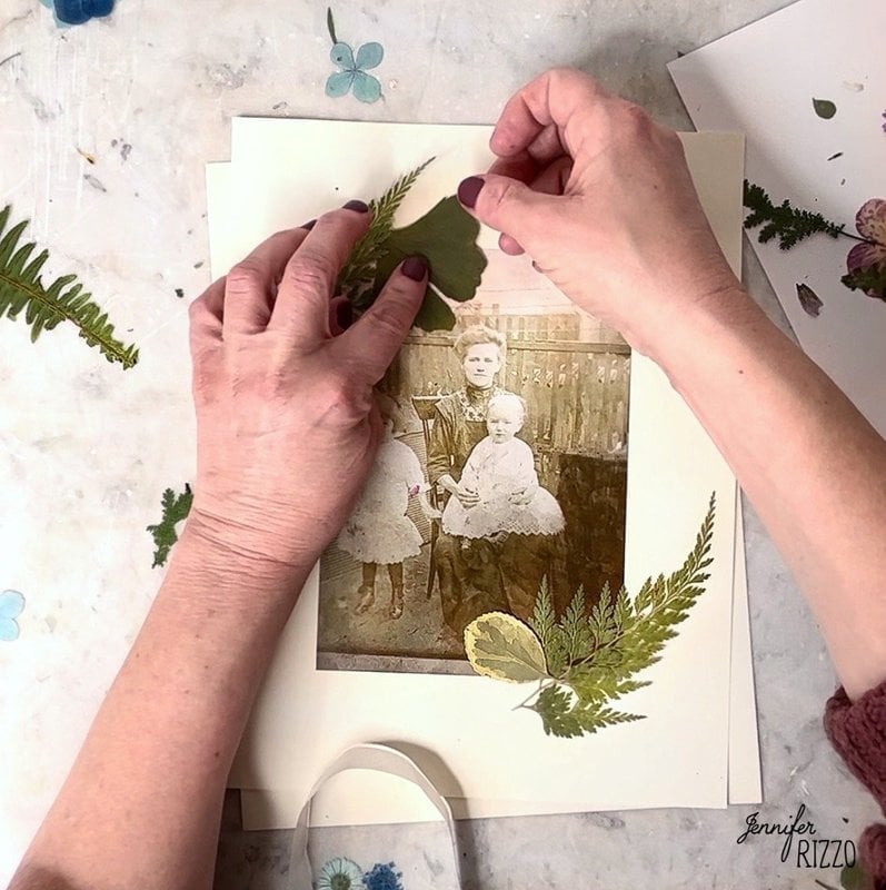 Hands arranging pressed leaves and ferns around a vintage sepia photograph of three people on a light surface, creating the perfect base to make a framed dried flower vintage photo collage.