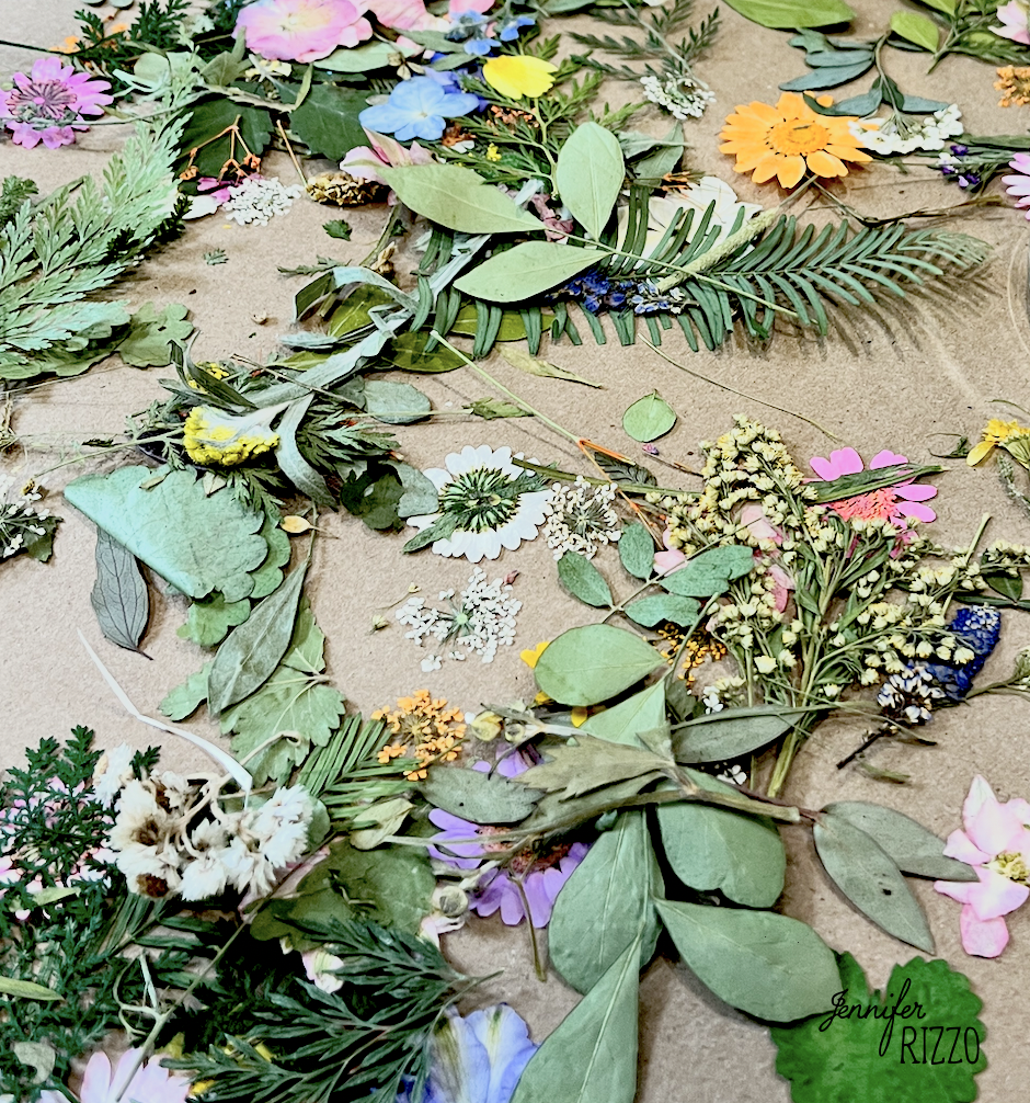 Assorted dried leaves, flowers, and greenery are scattered on a brown surface, displaying a variety of textures and colors—perfect materials to make a framed dried flower vintage photo collage.