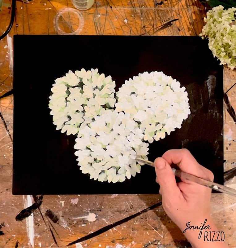 A hand paints white hydrangea flowers on a black canvas. Art supplies and flowers are scattered on a wooden table.