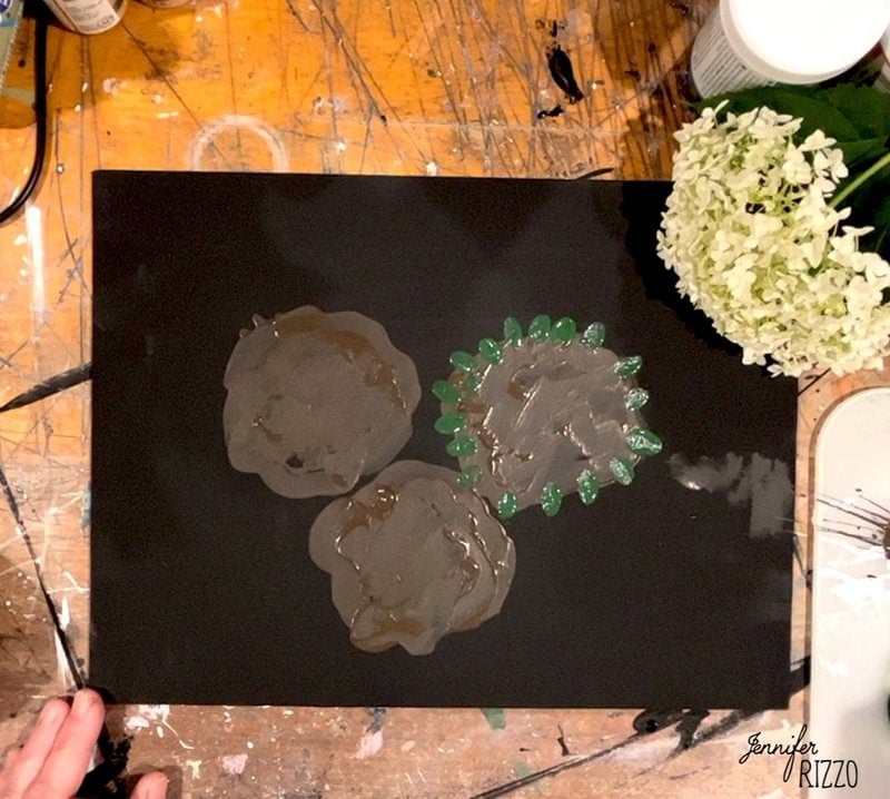 A black sheet with three blobs of wet gray paint, one edged with green dots, on a messy wooden table beside white hydrangea flowers.