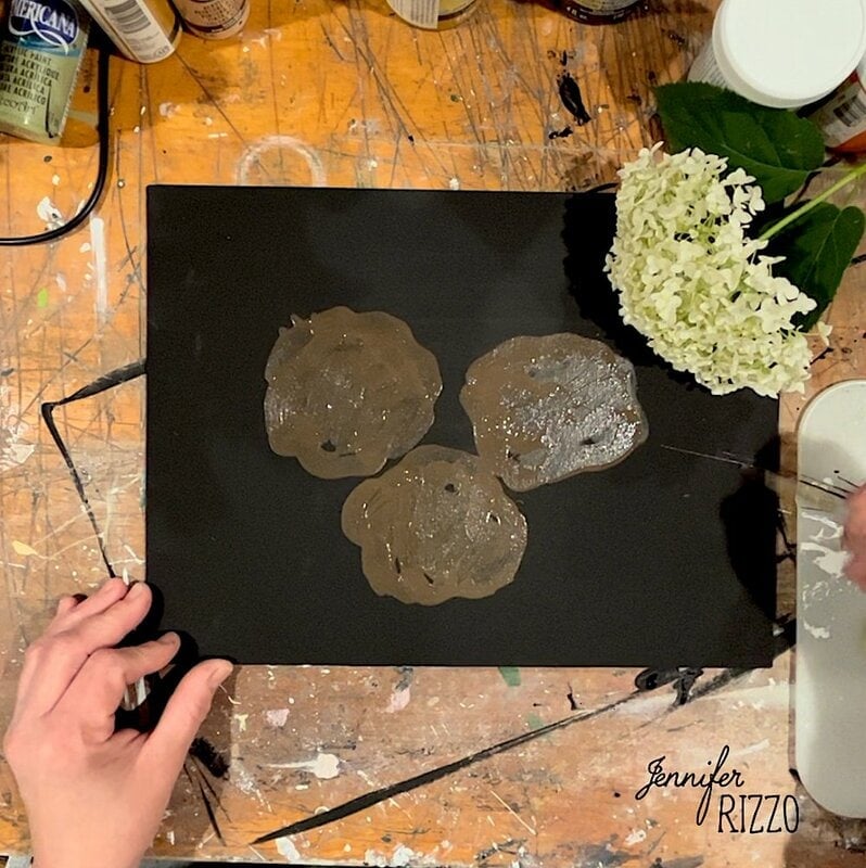 Hands working on a black canvas with three brown paint blobs, surrounded by art supplies and a white hydrangea on a messy table.