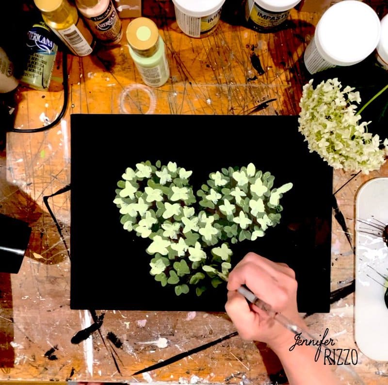 A person paints green and yellow leaves in the shape of a heart on a black canvas, surrounded by art supplies on a messy wooden table.