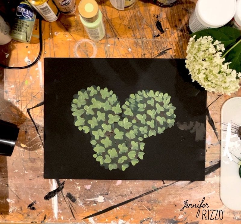 A painting of green leaves arranged in the shape of Mickey Mouse on black paper, surrounded by art supplies and a white flower on a wooden table.