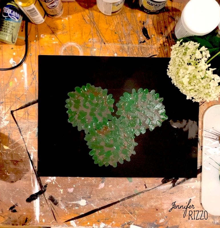 Three green painted leaves on black paper, surrounded by art supplies, a white flower, and a wooden table marked with paint stains.