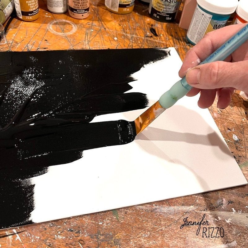 A hand uses a paintbrush to spread black paint across a blank canvas on a cluttered wooden table with paint bottles.