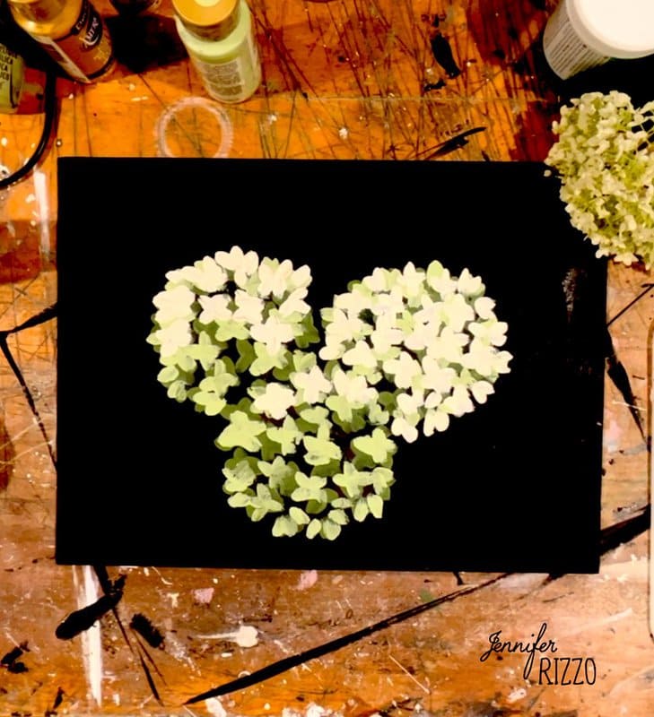 A heart shape made of green hydrangea petals is arranged on a black canvas, surrounded by art supplies on a paint-stained wooden table.