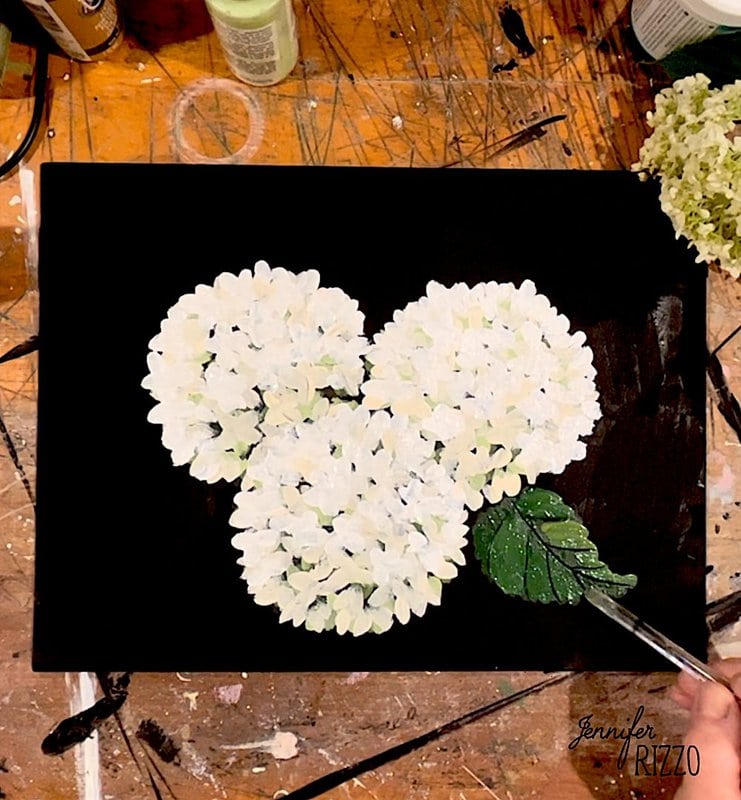A hand paints white hydrangea flowers and a green leaf on a black canvas, surrounded by art supplies on a wooden table.
