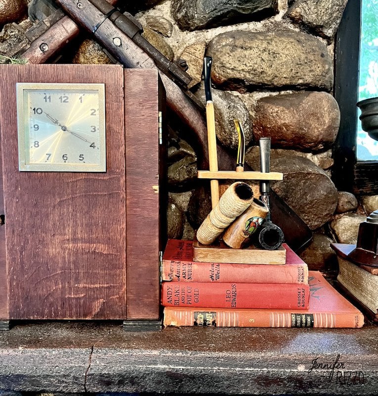 A Camp Wandawega Vintage Decor touch: a wooden clock sits next to pipes in a stand atop four red hardcover books, set before a stone wall.
