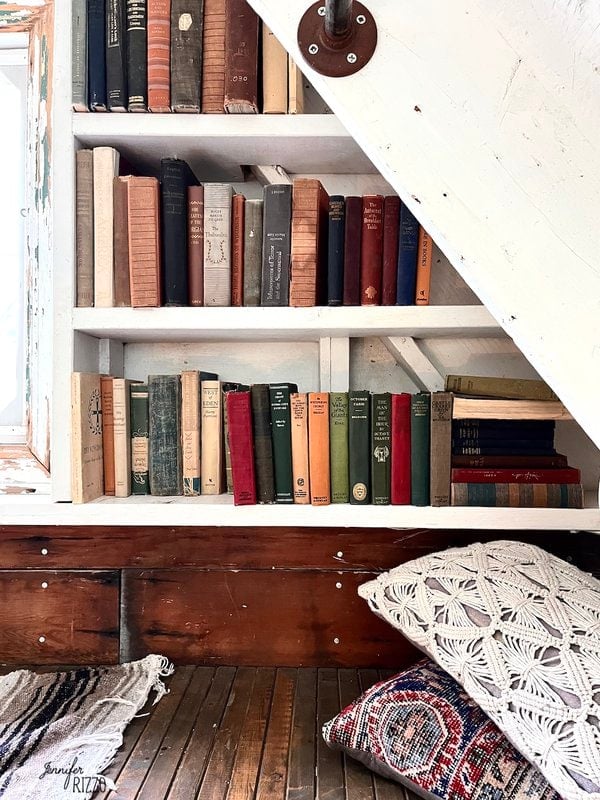 A built-in bookshelf under a white staircase, inspired by Camp Wandawega Vintage Decor, holds assorted vintage books; below, a patterned rug and two textured pillows are placed on a wooden bench.