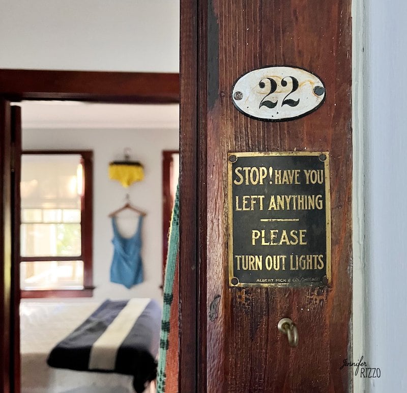 Close-up of a door with room number 22, featuring Camp Wandawega vintage decor and a sign reminding guests to check for belongings and turn out lights; a bed and clothing are visible in the room beyond.