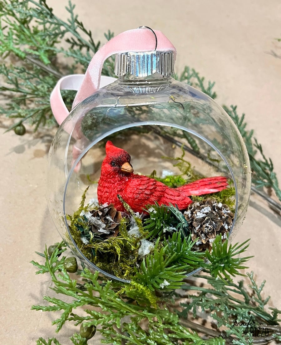 A clear ornament, inspired by DIY hanging snow globe ornaments, contains a red cardinal figurine, faux moss, and pinecones, with a pink ribbon for hanging. Greenery surrounds the ornament.