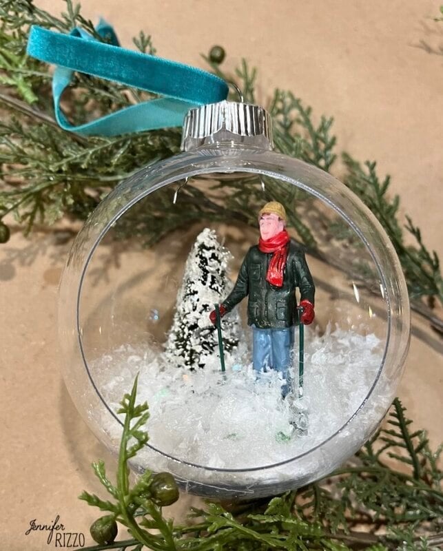 A clear Christmas ornament crafted as a DIY hanging snow globe features a figurine in winter clothing with ski poles, standing on artificial snow beside a snow-covered tree.
