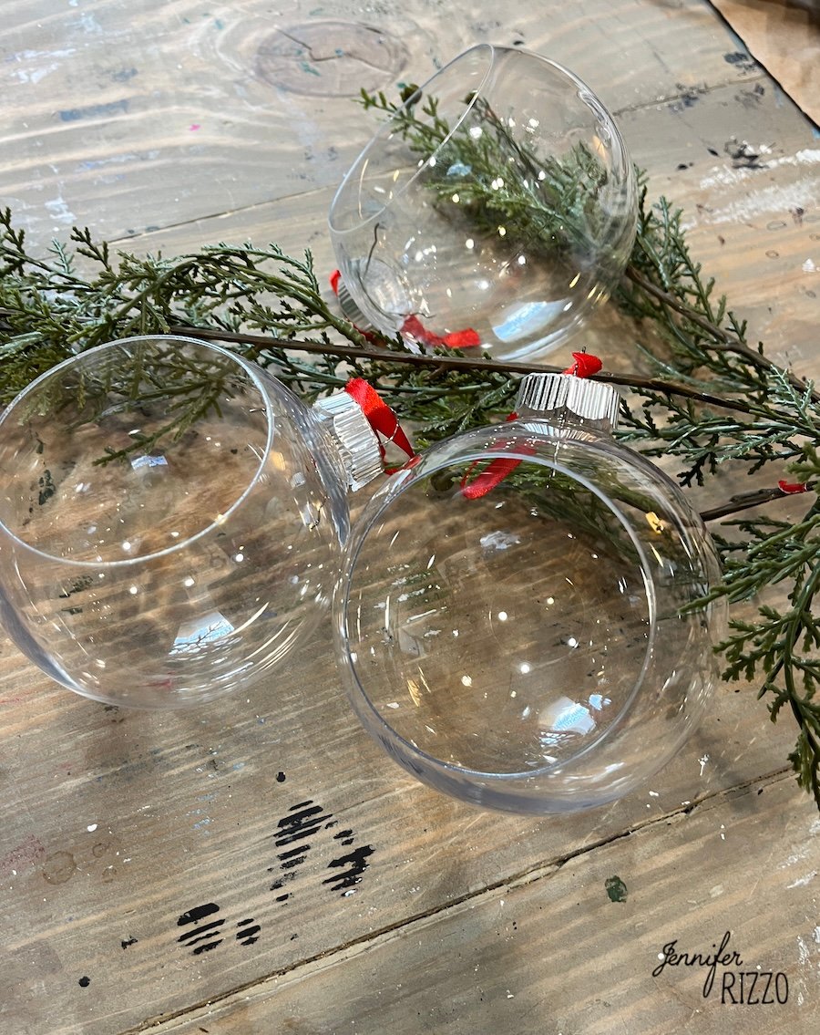 Three empty clear glass ornaments rest on a wooden surface next to sprigs of greenery with small red berries, ready to be transformed into festive DIY hanging snow globe ornaments.