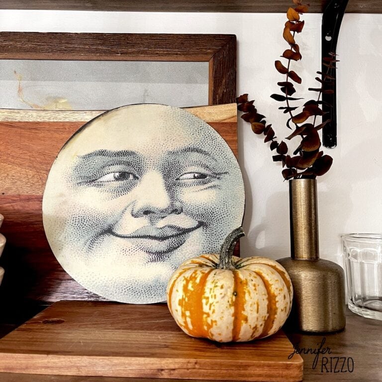 Halloween Man in the moon face shelf decor with a mini pumpkin and a brass vase with eucalyptus stems some out of it sitting on a cutting board