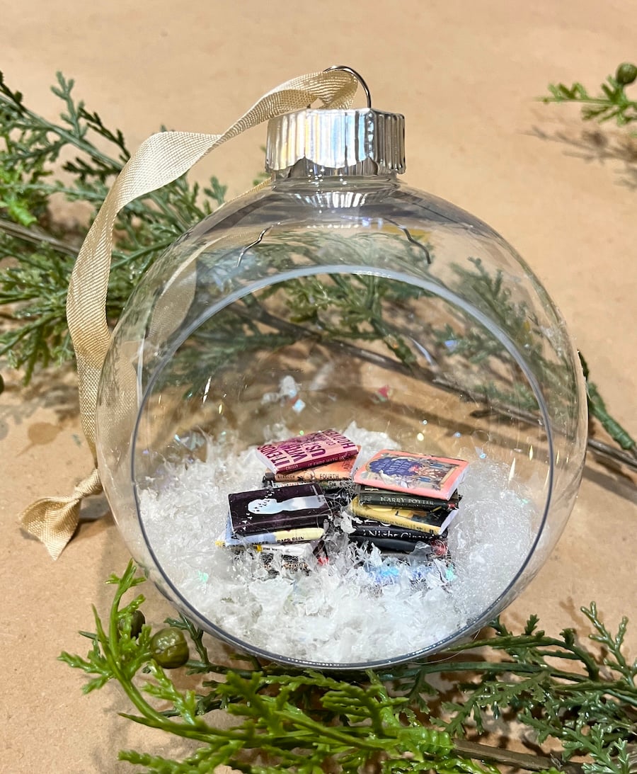 A clear glass DIY hanging snow globe ornament filled with artificial snow and miniature books, suspended from a gold ribbon, with greenery in the background.