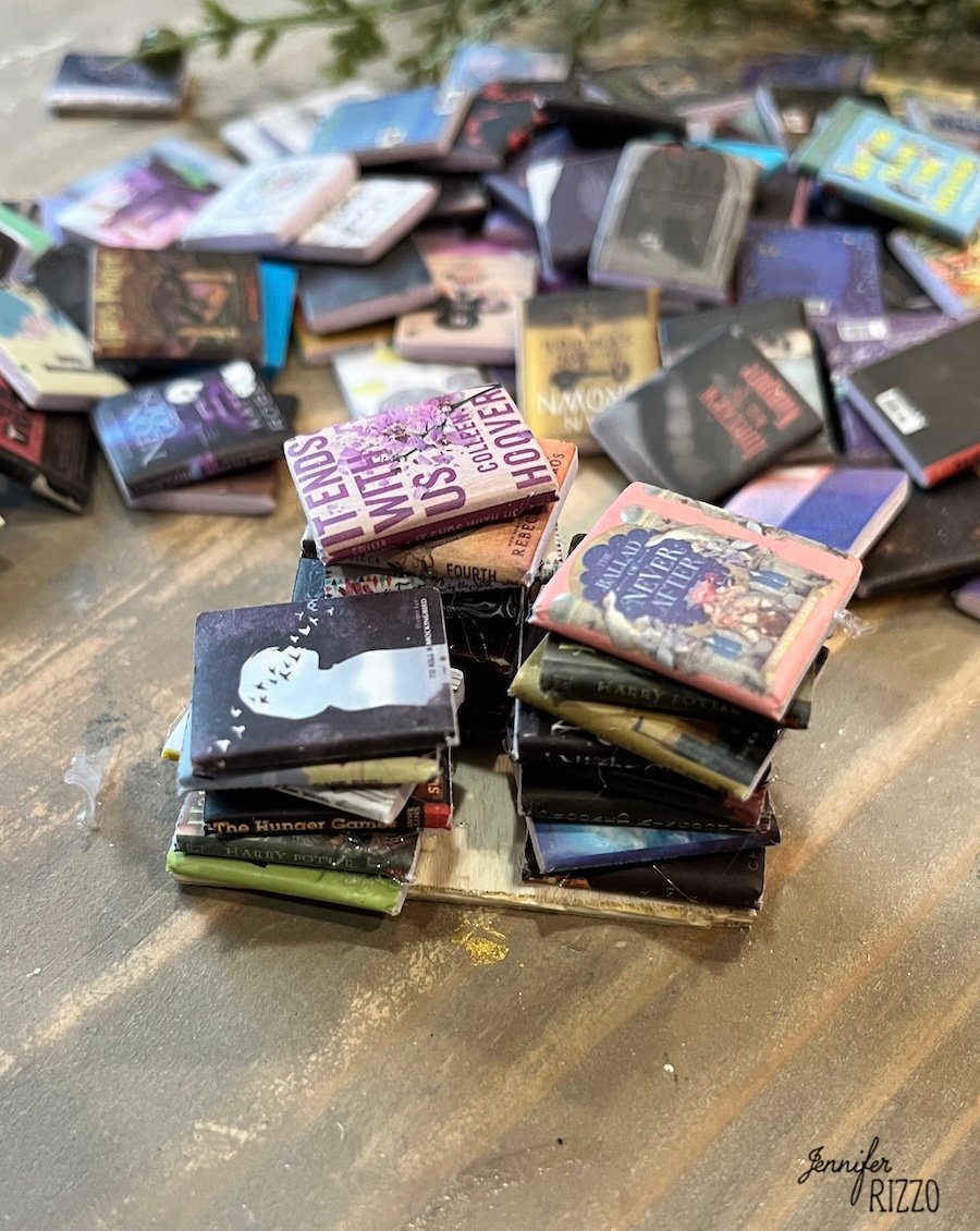 Stacks of miniature books are arranged on a wooden surface, with more scattered in the background. The colorful covers create a cozy scene, perfect for pairing with DIY hanging snow globe ornaments for festive decor.