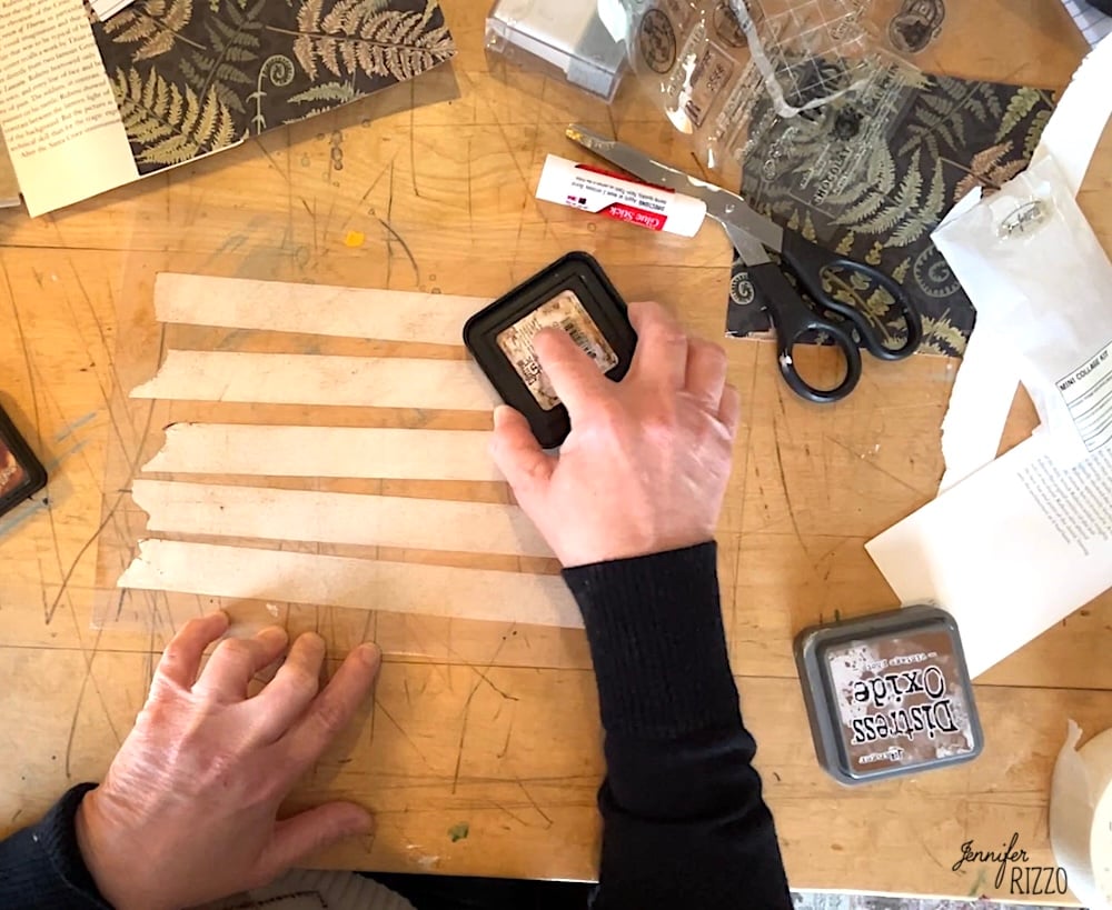 A person uses a Distress Oxide ink pad on strips of paper at a cluttered craft table, demonstrating How to Make Your Own Custom DIY Washi Tape with scissors, glue stick, stamps, and patterned paper.