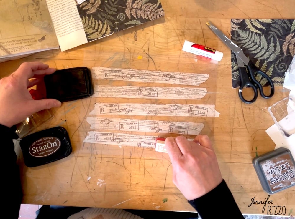 Person crafting at a table, learning how to make your own custom DIY washi tape by applying stamps to paper strips with an ink pad and glue stick. Scissors, papers, and crafting supplies are scattered around.