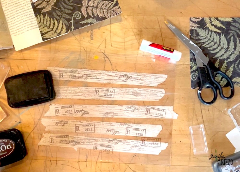 A table with strips of patterned paper, a glue stick, scissors, ink pads, cut paper pieces, and a decorative fern-patterned sheet—perfect supplies for learning how to make your own custom DIY washi tape.