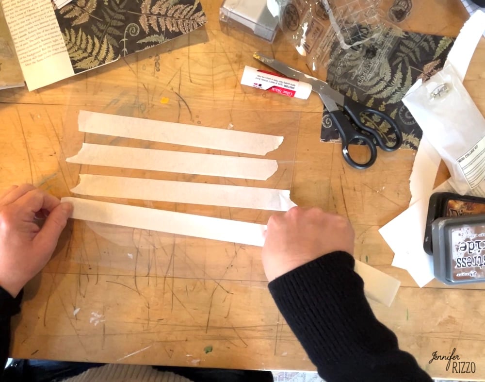 Person tearing strips of white paper on a wooden desk cluttered with scissors, a glue stick, ink pads, stamps, and patterned paper—gathering supplies for How to Make Your Own Custom DIY Washi Tape.