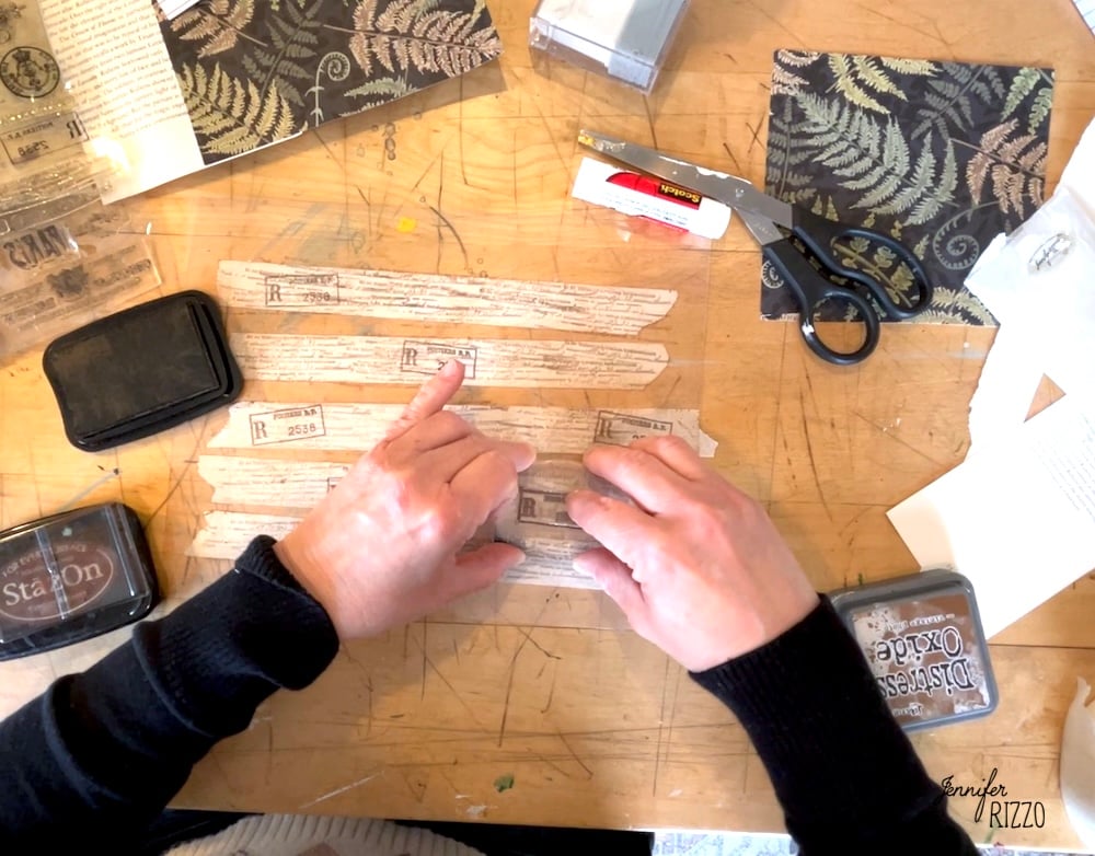 A person arranges strips of printed tape on a wooden table, surrounded by crafting supplies like scissors, glue, ink pads, and decorative paper—perfect for learning how to make your own custom DIY washi tape.