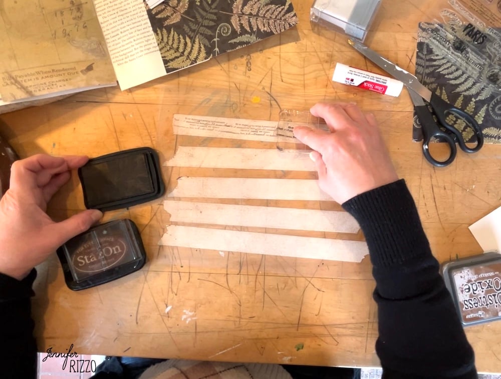 Person using a black ink pad and clear stamp on strips of paper at a cluttered craft table, creating designs for How to Make Your Own Custom DIY Washi Tape with scissors, glue stick, and decorative papers.