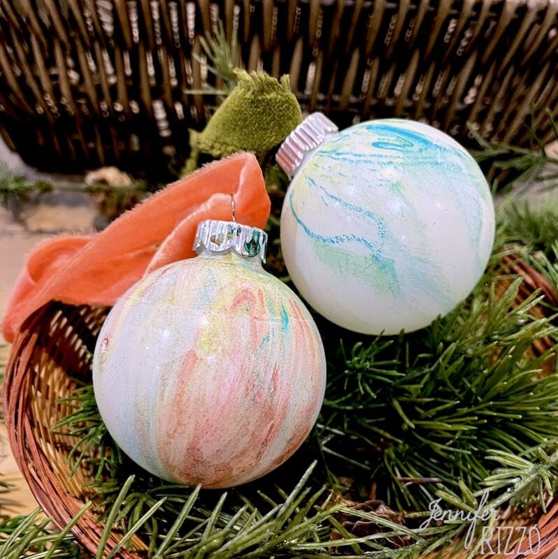 Two marble-patterned Christmas ornaments rest on pine branches in a woven basket, one with a red ribbon attached—an inspiring example of how to make marbled Christmas ornaments with paint and pastel swirls.