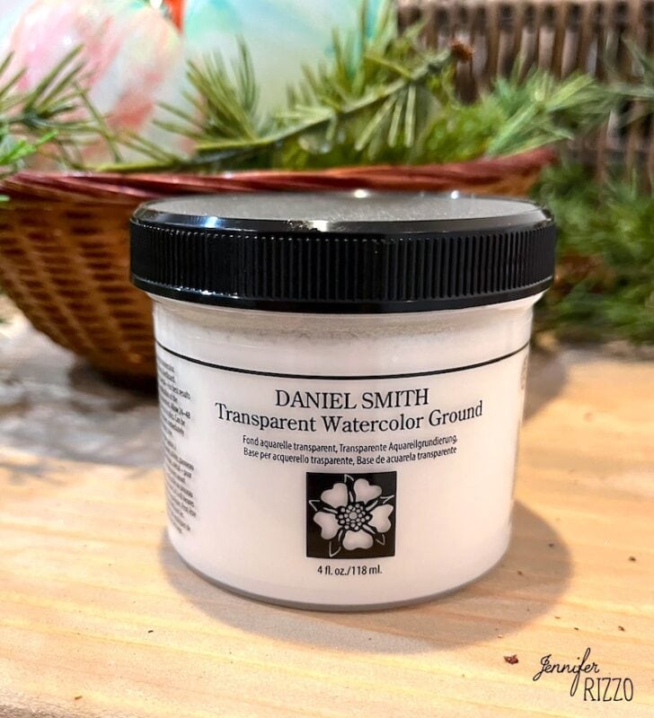 A jar of Daniel Smith Transparent Watercolor Ground sits on a wooden surface, perfect for learning how to make marbled Christmas ornaments with paint. Greenery and a basket are blurred in the background, creating a festive atmosphere.