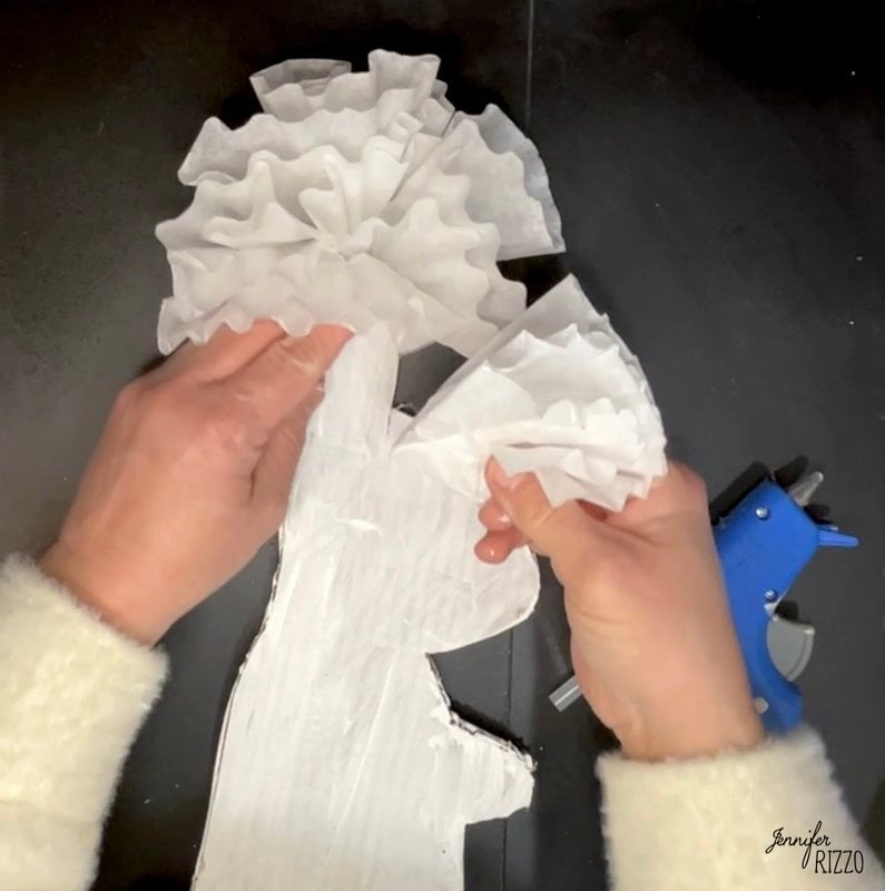 Two hands attach white coffee filters to a white-painted cardboard bunny cutout, with a blue hot glue gun nearby on a black surface in this fun Coffee Filter Easter Bunny Craft.