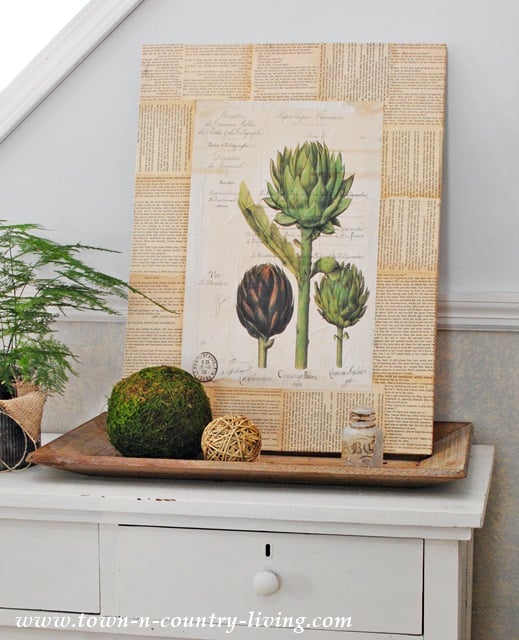A framed DIY botanical art print with book page borders sits on a white dresser, next to a potted fern, moss ball, and decorative twine balls arranged on a wooden tray.