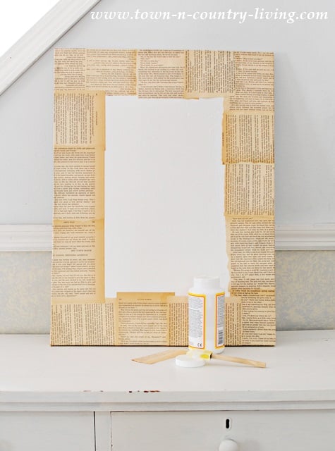A mirror with a frame decorated using vintage book pages and touches of DIY botanical art sits on a white table, with a bottle of glue and a brush placed in front of it.