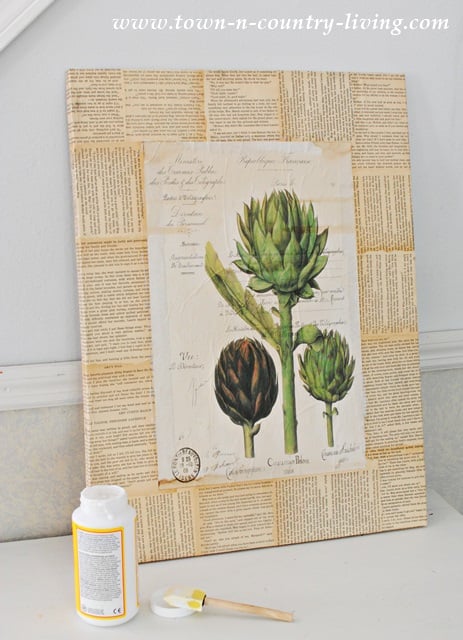 A canvas decorated with vintage book pages and an artichoke botanical print—perfect for DIY botanical art—is propped against a wall; a plastic glue bottle, sponge brush, and lid sit on the table in front.