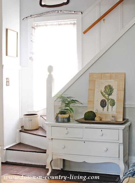 A white dresser sits by a staircase with a potted plant, DIY botanical art featuring a framed artichoke print, and decorative items on top; sunlight filters through a window beside the stairs.
