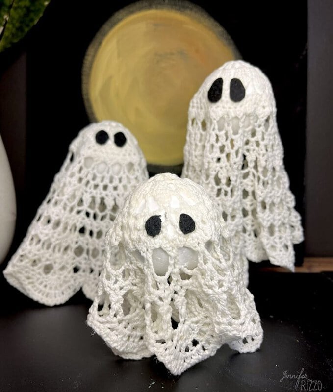 Three small ghost decorations made from white crochet doilies with black eyes, arranged in front of a round yellow and brown background—perfect for adding a whimsical touch to your fall decor projects.