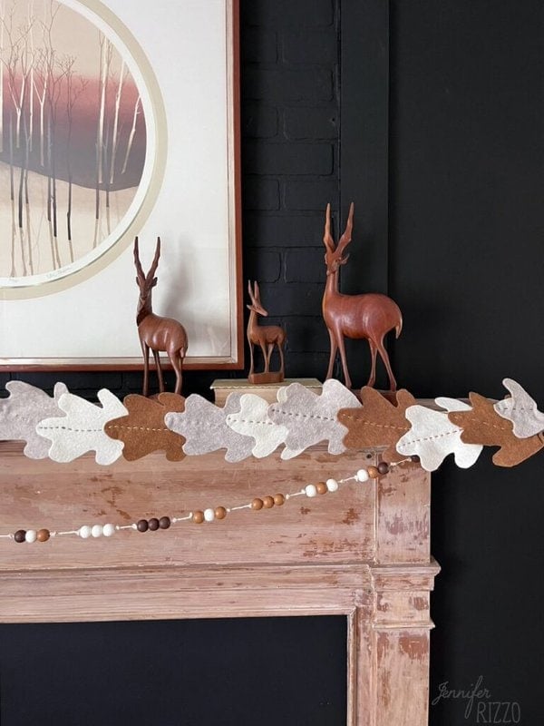A wooden fireplace mantel with three deer figurines and oak leaf decorations. A circular framed artwork hangs above the mantel. A garland with beads and felt leaves is draped across the mantel from Jenniferrizzo.cm