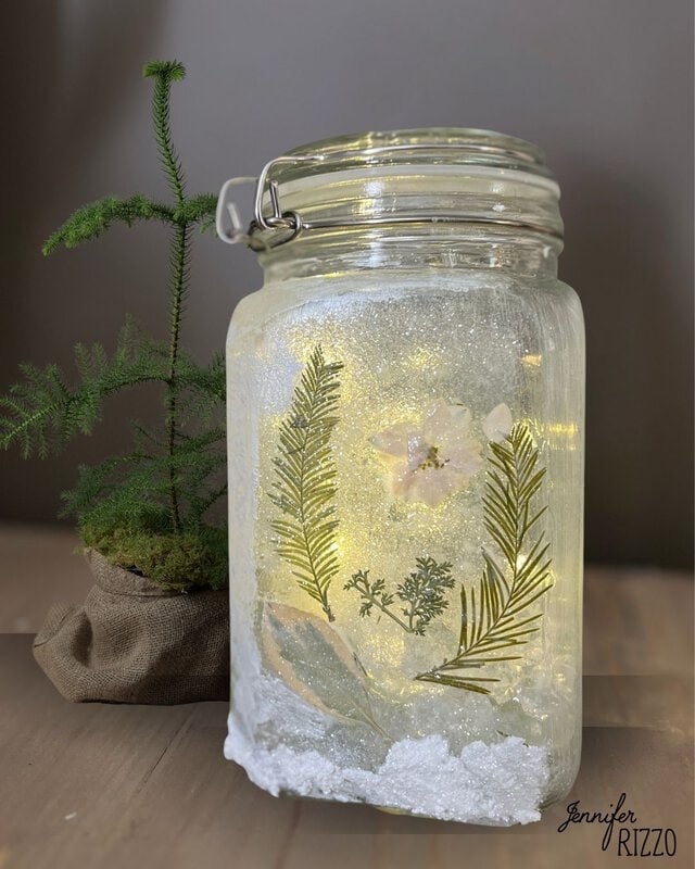 A glass jar with embedded flowers and leaves is illuminated from within, next to a small potted plant.