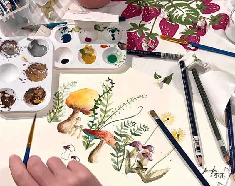 A hand painting watercolor mushrooms and plants on paper, surrounded by paint palettes, brushes, and colored pencils.