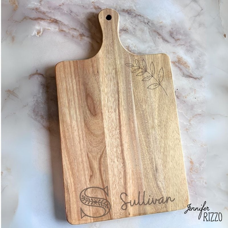 Wooden cutting board with a handle, engraved with the name "Sullivan" and a leaf design. Signature "Jennifer Rizzo" at the bottom right.