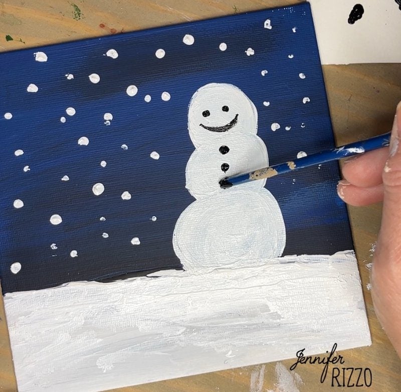 Hand painting a smiling snowman on a square canvas with falling snow against a dark blue background.